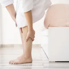 Une personne en peignoir blanc se tient près d'un lit, se massant la jambe. Elle cherche des remèdes de grand-mère pour ses jambes lourdes.
