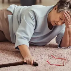 Une femme âgée est tombée et peine à se relever, sa canne et ses lunettes à portée de main. Elle manque de plus de force dans les jambes pour se relever.