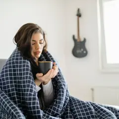 Femme emmitouflée dans une couverture, buvant une tasse chaude, ressentant des frissons sans fièvre. Une guitare est accrochée au mur.