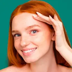 Une jeune femme rousse souriante, peau parsemée de taches de rousseur, touche son front. Elle utilise peut-être un soin au zinc pour la peau.