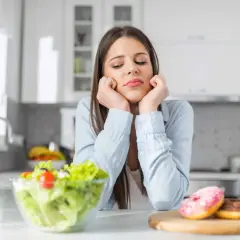Une jeune femme dépitée face à une salade et des donuts, symbolisant le dilemme des aliments à éviter pour la dermite séborrhéique.