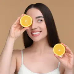 Une femme souriante tient des moitiés d'orange devant ses yeux. Attention aux aliments acides à éviter pour une peau éclatante.