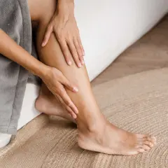 Une personne se tient la jambe gauche, peut-être à cause d'une douleur jambe gauche. Elle est assise sur un lit, enveloppée dans un peignoir gris.