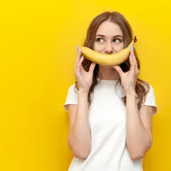 Une jeune femme tient une banane devant sa bouche, souriant. La question "la banane fait elle grossir ?" flotte dans l'air.