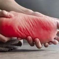 Main gauche, la plante du pied est tenue et massée, avec une zone rouge vif indiquant une douleur au pied gauche.