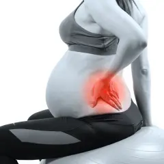 Femme enceinte assise sur un ballon de gym, main sur le bas du dos, zone rouge indiquant une douleur au bassin pendant la grossesse.