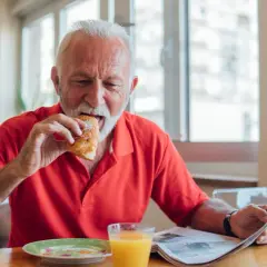 Un homme âgé mange un croissant en lisant le journal, un verre de jus d'orange à côté. Une scène qui illustre les besoins nutritionnels de la personne âgée.