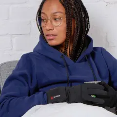 Femme avec des tresses, lunettes, sweat bleu et gants chauffants G-HEAT, assise sur un canapé, elle semble avoir toujours froid.