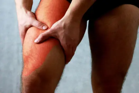 Main gauche tenant la cuisse droite, marquée d'une zone rouge symbolisant une douleur haut de la cuisse.