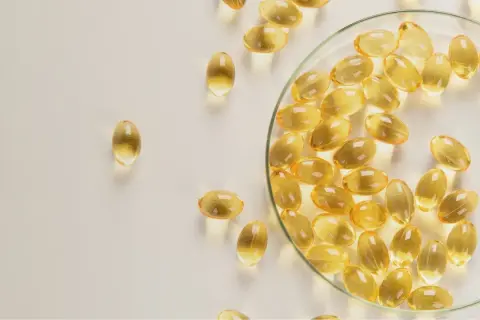 Gélules d'huile de poisson oméga 3 translucides, certaines dans une coupelle en verre, sur fond clair.