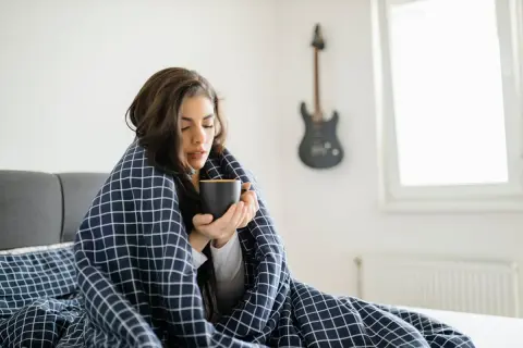 Femme emmitouflée dans une couverture, buvant une tasse chaude, ressentant des frissons sans fièvre. Une guitare est accrochée au mur.