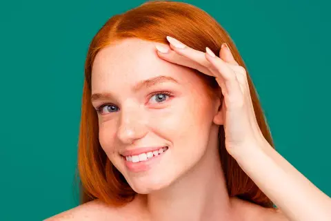 Une jeune femme rousse souriante, peau parsemée de taches de rousseur, touche son front. Elle utilise peut-être un soin au zinc pour la peau.