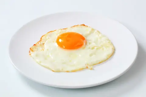 Un œuf au plat, bien cuit, sur une assiette blanche. Le jaune est bien rond et brillant, parfait pour un petit-déjeuner nourrissant.