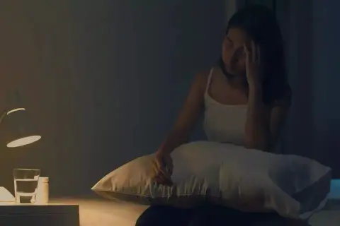 Femme insomniaque, le front serré, envisageant une cure de magnésium. Sur la table de nuit, une lampe, une bouteille d'eau et des médicaments.