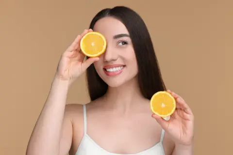 Une femme souriante tient des moitiés d'orange devant ses yeux. Attention aux aliments acides à éviter pour une peau éclatante.