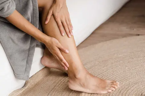 Une personne se tient la jambe gauche, peut-être à cause d'une douleur jambe gauche. Elle est assise sur un lit, enveloppée dans un peignoir gris.