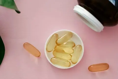 Gélules de collagène et vitamine C sur fond rose, à côté d'une feuille verte.