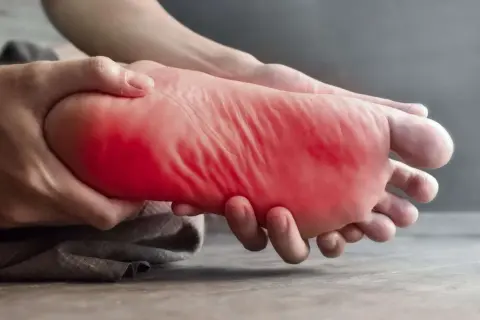 Main gauche, la plante du pied est tenue et massée, avec une zone rouge vif indiquant une douleur au pied gauche.