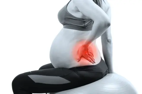 Femme enceinte assise sur un ballon de gym, main sur le bas du dos, zone rouge indiquant une douleur au bassin pendant la grossesse.