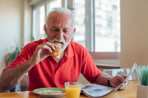 Un homme âgé mange un croissant en lisant le journal, un verre de jus d'orange à côté. Une scène qui illustre les besoins nutritionnels de la personne âgée.