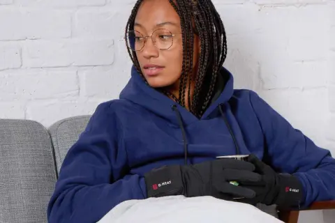 Femme avec des tresses, lunettes, sweat bleu et gants chauffants G-HEAT, assise sur un canapé, elle semble avoir toujours froid.