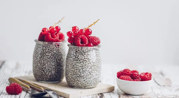 Deux pots de crème de chia garnis de framboises et de groseilles. Un petit bol de framboises à côté. Parfait pour une graine de chia enceinte.