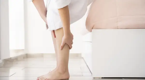 Une personne en peignoir blanc se tient près d'un lit, se massant la jambe. Elle cherche des remèdes de grand-mère pour ses jambes lourdes.