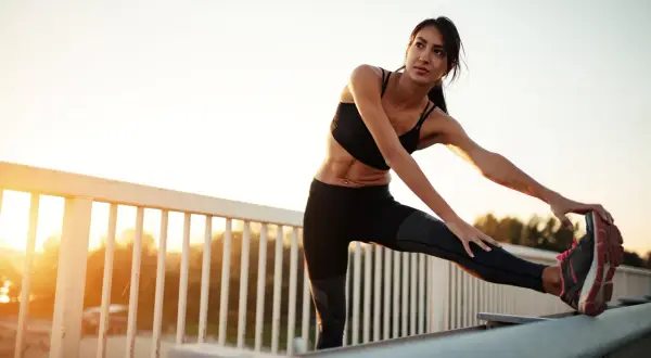 Femme s'étirant les ischio-jambiers, une conséquence d'un entraînement intense. Le soleil se lève sur un paysage flou.