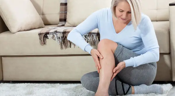 Femme assise sur un tapis blanc, touchant sa jambe. Elle ressent une douleur derrière la cuisse sans effort, visiblement préoccupée.