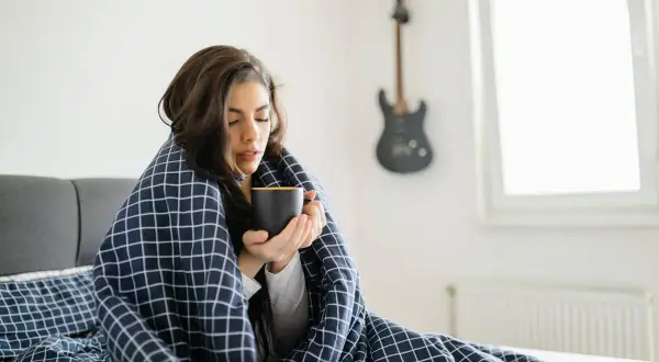 Femme emmitouflée dans une couverture, buvant une tasse chaude, ressentant des frissons sans fièvre. Une guitare est accrochée au mur.