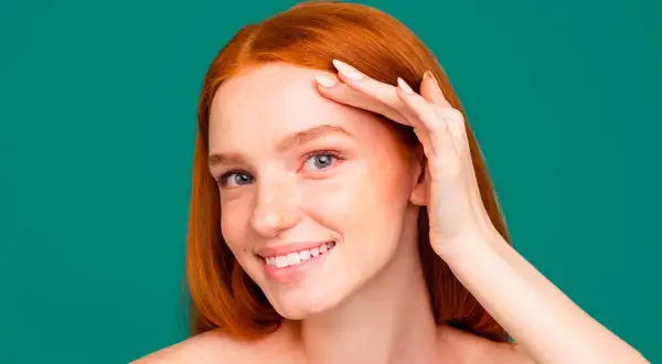 Une jeune femme rousse souriante, peau parsemée de taches de rousseur, touche son front. Elle utilise peut-être un soin au zinc pour la peau.