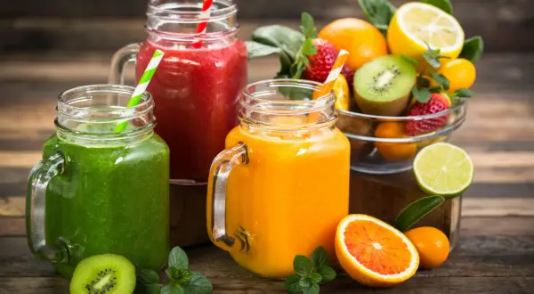 Trois smoothies colorés (vert, rouge, orange) dans des bocaux en verre, accompagnés de fruits frais. Parfait pour une cure detox maison.