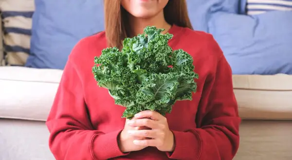 Une femme souriante tient un bouquet de chou kale, un légume aux multiples chou kale bienfaits pour la santé.