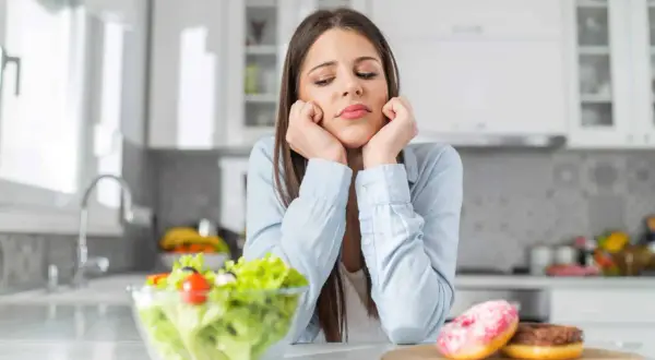 Une jeune femme dépitée face à une salade et des donuts, symbolisant le dilemme des aliments à éviter pour la dermite séborrhéique.