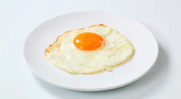 Un œuf au plat, bien cuit, sur une assiette blanche. Le jaune est bien rond et brillant, parfait pour un petit-déjeuner nourrissant.