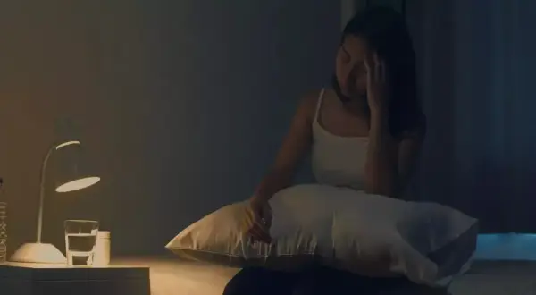 Femme insomniaque, le front serré, envisageant une cure de magnésium. Sur la table de nuit, une lampe, une bouteille d'eau et des médicaments.