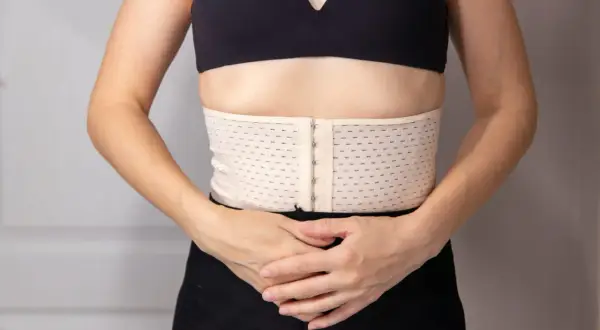Ceinture de resserrage du bassin, beige, avec fermeture à crochets, portée sous un soutien-gorge noir.