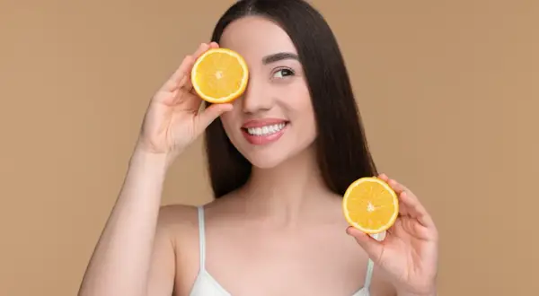 Une femme souriante tient des moitiés d'orange devant ses yeux. Attention aux aliments acides à éviter pour une peau éclatante.
