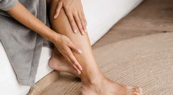 Une personne se tient la jambe gauche, peut-être à cause d'une douleur jambe gauche. Elle est assise sur un lit, enveloppée dans un peignoir gris.