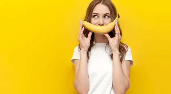 Une jeune femme tient une banane devant sa bouche, souriant. La question "la banane fait elle grossir ?" flotte dans l'air.