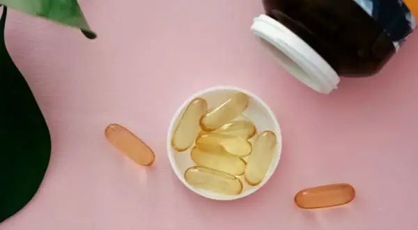 Gélules de collagène et vitamine C sur fond rose, à côté d'une feuille verte.