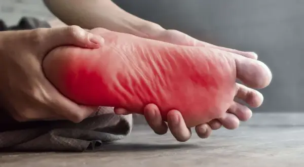 Main gauche, la plante du pied est tenue et massée, avec une zone rouge vif indiquant une douleur au pied gauche.