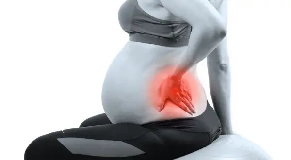 Femme enceinte assise sur un ballon de gym, main sur le bas du dos, zone rouge indiquant une douleur au bassin pendant la grossesse.