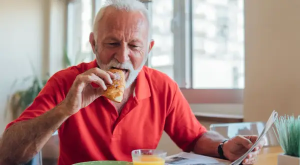 Un homme âgé mange un croissant en lisant le journal, un verre de jus d'orange à côté. Une scène qui illustre les besoins nutritionnels de la personne âgée.