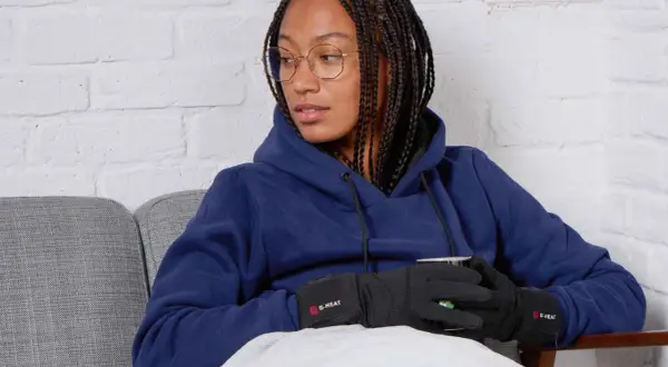 Femme avec des tresses, lunettes, sweat bleu et gants chauffants G-HEAT, assise sur un canapé, elle semble avoir toujours froid.