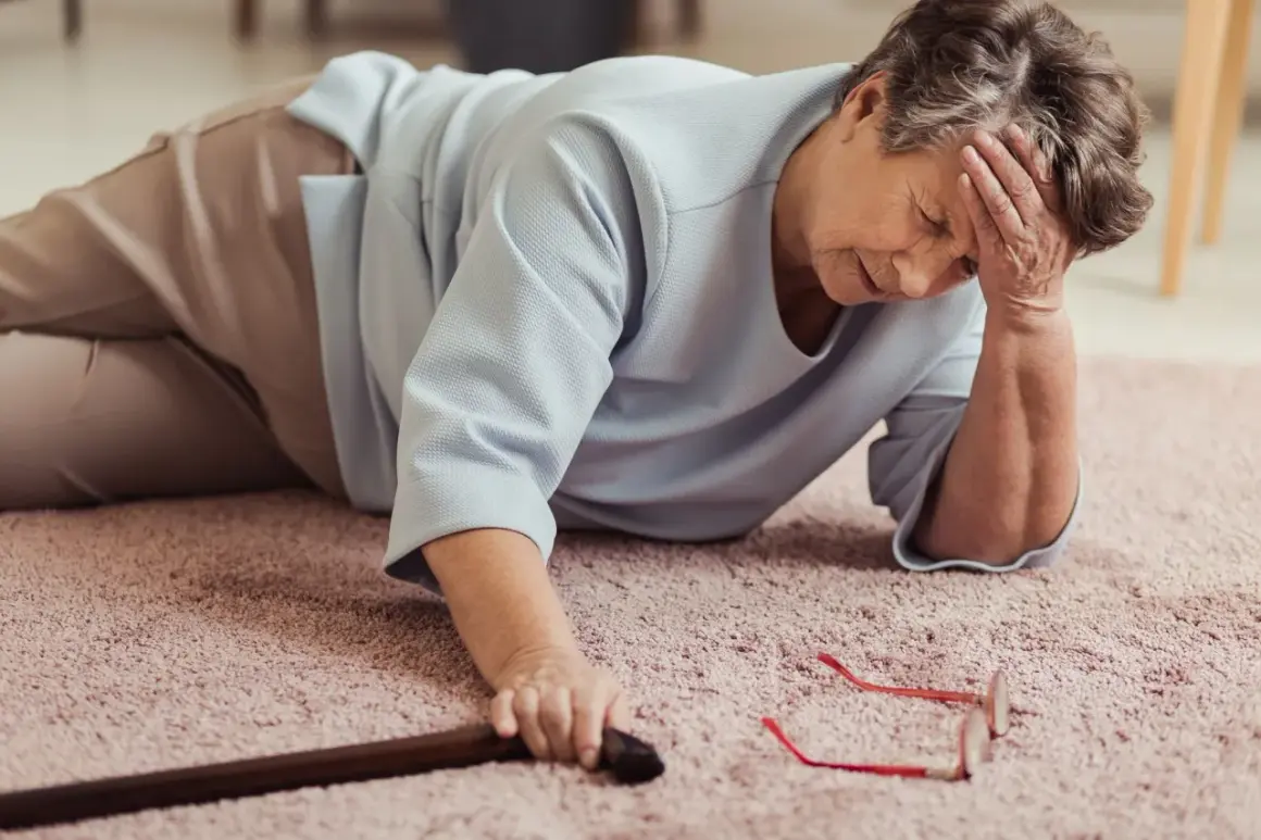 Une femme âgée est tombée et peine à se relever, sa canne et ses lunettes à portée de main. Elle manque de plus de force dans les jambes pour se relever.