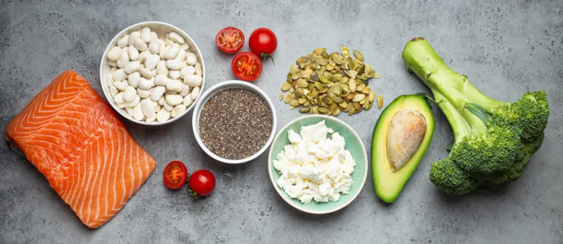 Saumon, haricots blancs, tomates, graines de chia, fromage blanc, avocat et brocoli sur fond gris.