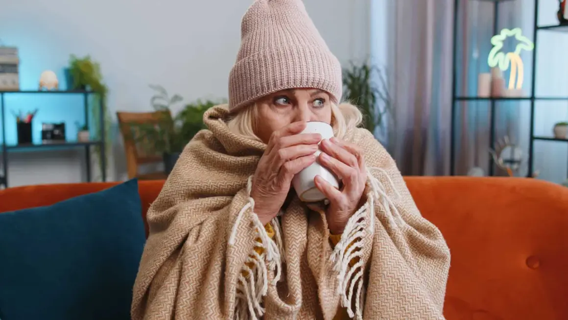 Une femme emmitouflée dans une couverture et un bonnet boit une tasse chaude, se demandant pourquoi j'ai toujours froid.