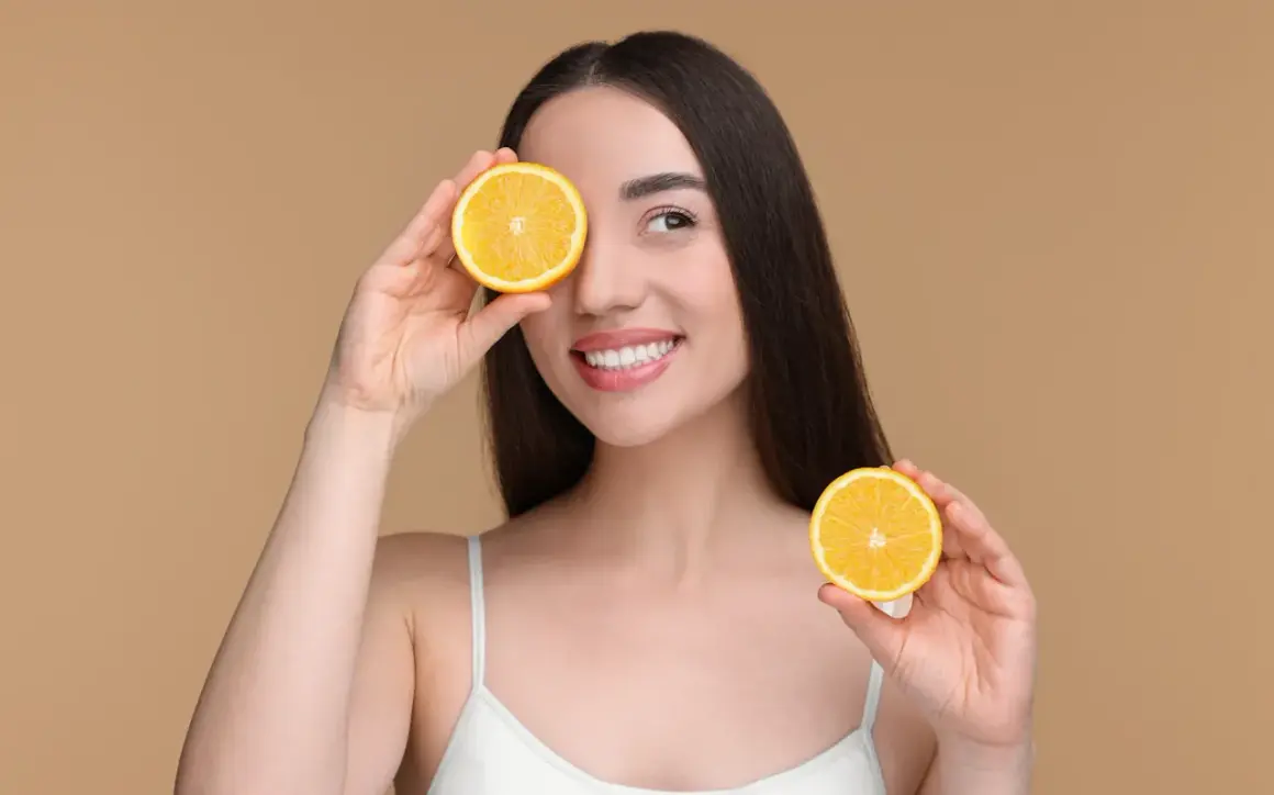 Une femme souriante tient des moitiés d'orange devant ses yeux. Attention aux aliments acides à éviter pour une peau éclatante.