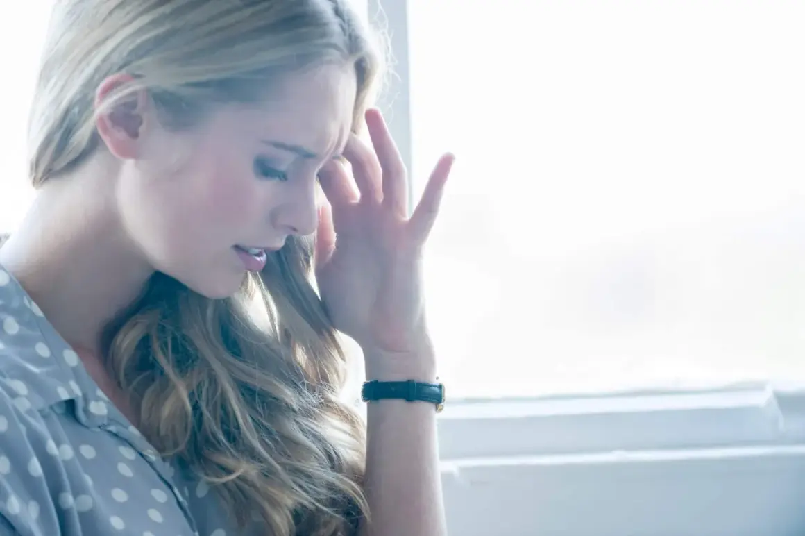 Femme blonde se tenant la tempe, le regard baissé, l'air de souffrir d'une sensation de pression dans la tête et vertige.